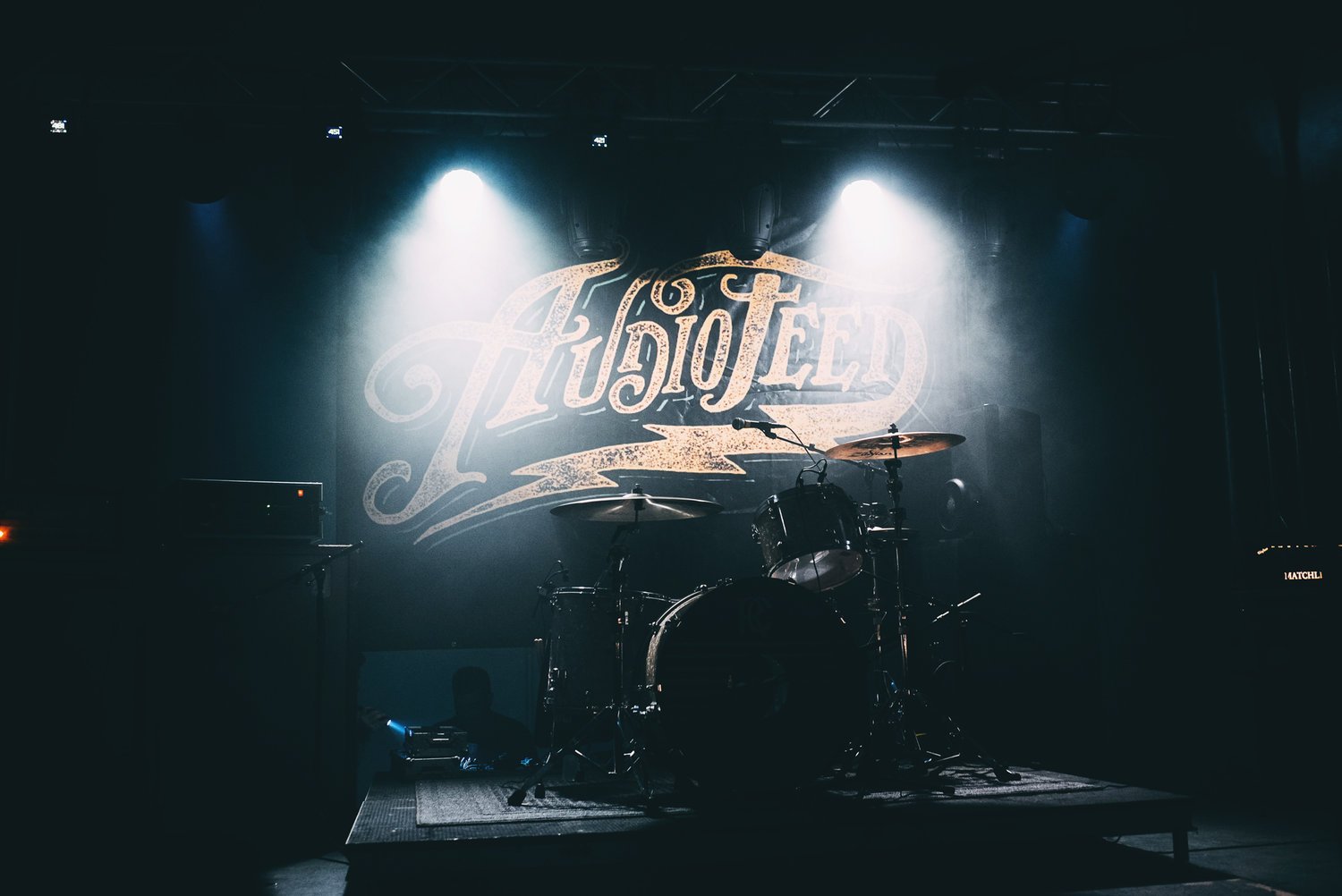 Photo of a drum set on a dimly lit stage with a banner that reads "Audiofeed" in the background.