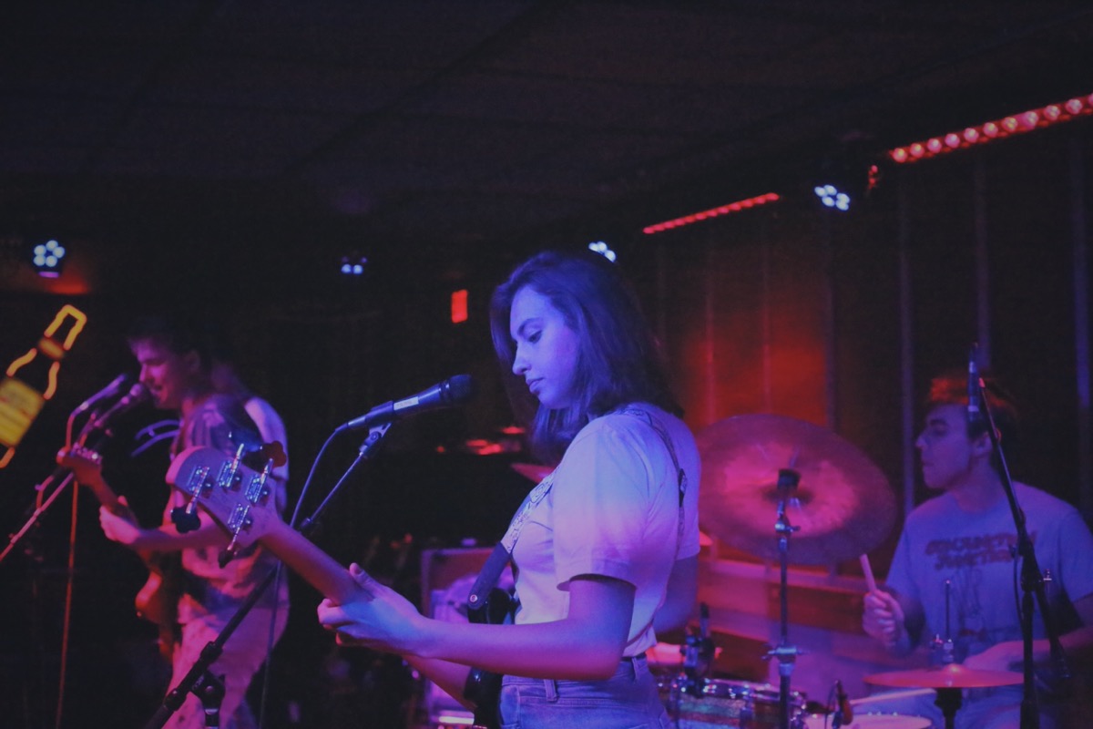 A musician playing bass in the foreground, a drummer and guitarist in the background