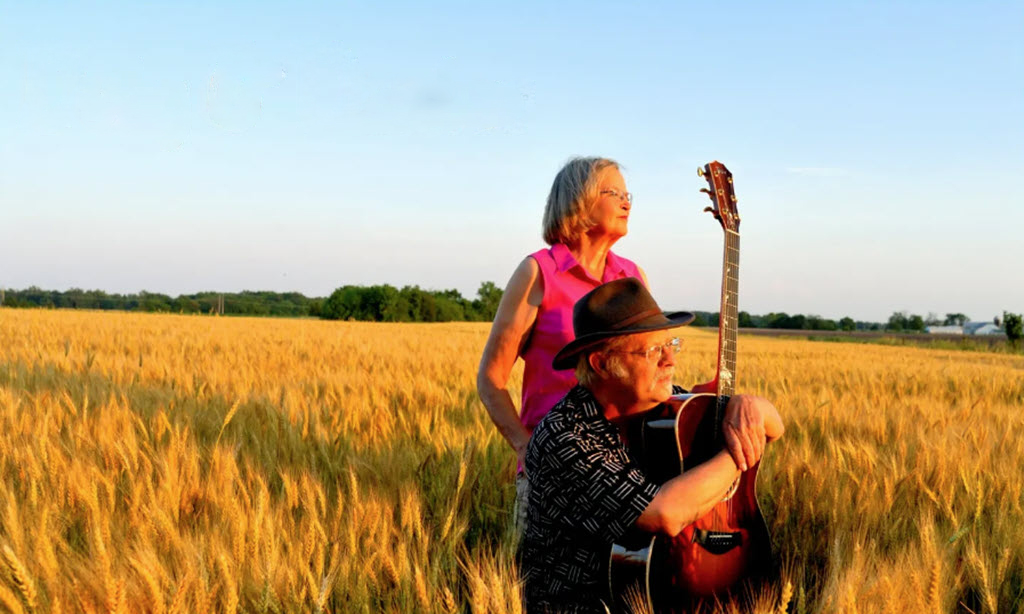 An older man and woman in a wheat field looking off into the distance. The man is kneeling and holding a guitar. The woman has a pink sleeveless blouse and is standing