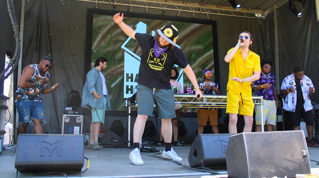 7 members of Half House performing onstage One member is dressed in birght yellow and has a microphone, another member is out front with a black hat with yellow flowers on it and another member is behind a table with DJ equipment.