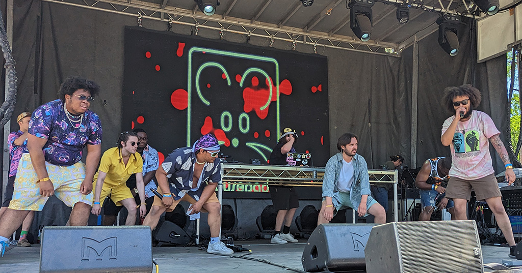 7 members of Half House on stage, hunched down as if they are swinging back and forth to the beat. One member has a microphone and another member is behind a table with DJ equipment.