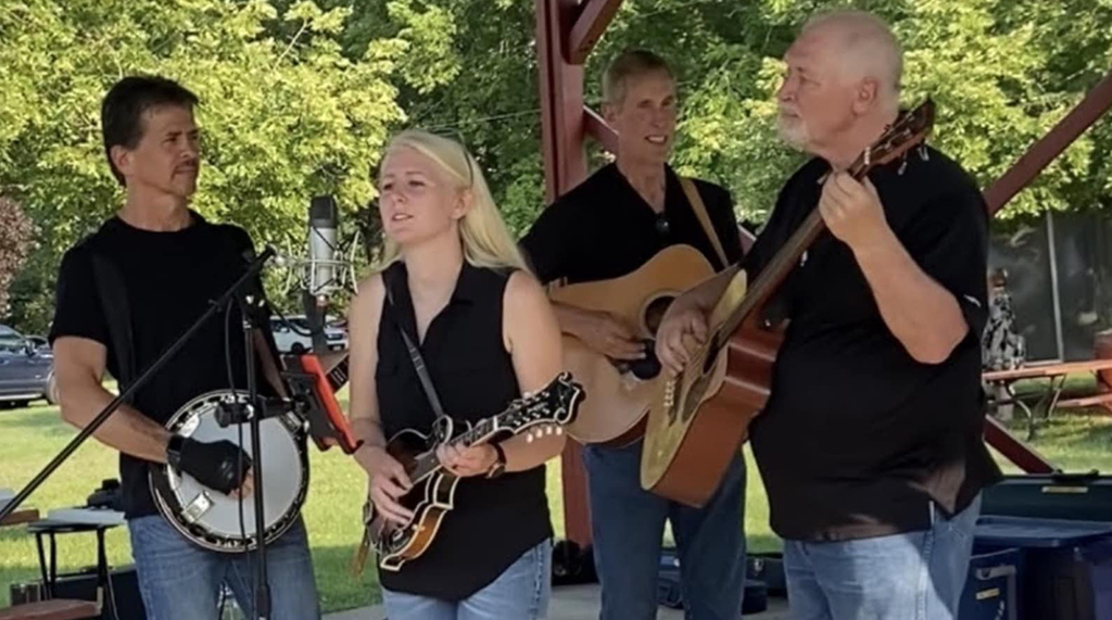 The four members of High Cotton Bluegrass Band onstage performing. 3 are male and one is female. They are all wearing black tops and blue jeans and are either playing guitar, banjo or mandolin.