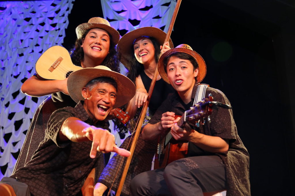 Honolulu theatre for youth. Two women stand smiling in back, with two men sitting in front. All are holding instruments, wearing straw hats, and smiling at the camera as they sing.