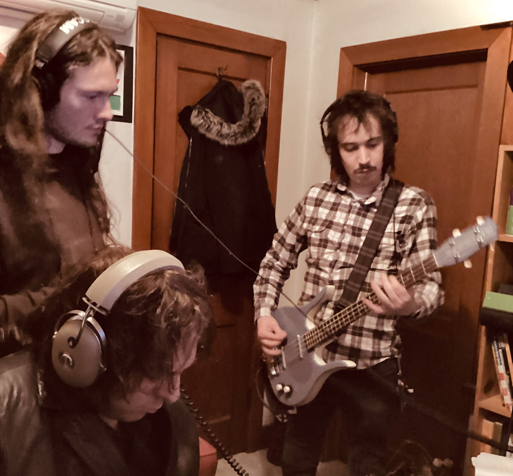 3 members of Mid Nite Leg recording in a small room. 3 members have headphones on, and one is playing the bass guitar.