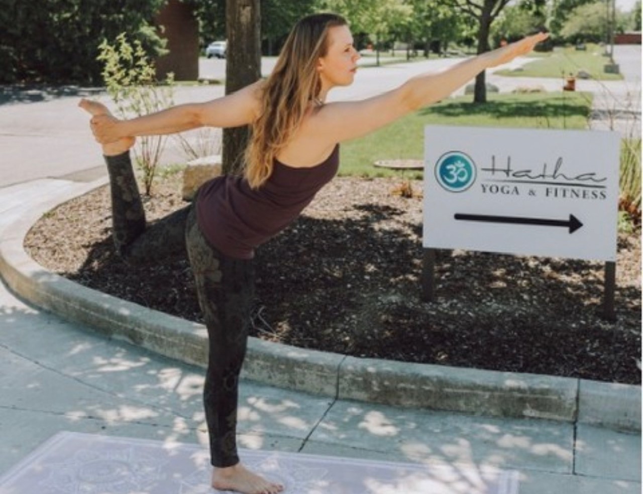 A white woman with long blonde hair is standing in a yoga pose with one arm outstretched in and the other holding her foot. She is standing on one leg on a yoga mat on an outdoor path.
