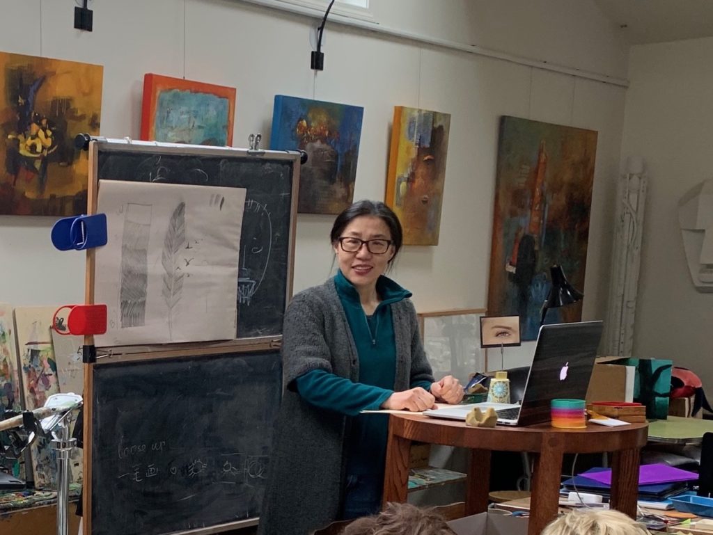 Hua Nian stands surrounded by paintings while teaching a class. She has dark hair pulled back, a turquoise long sleeve shirt and grey cardigan. She is looking and smiling at the camera