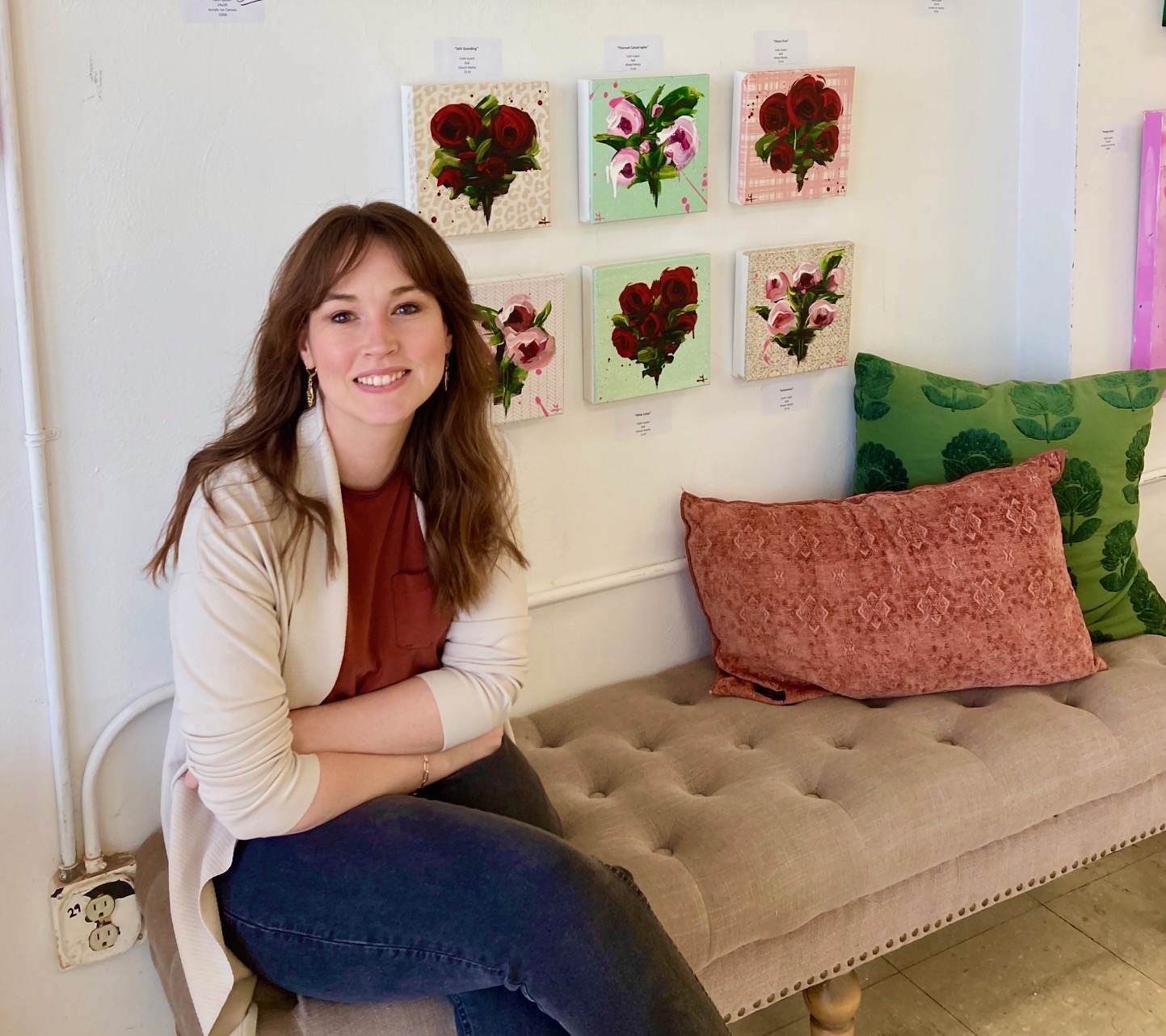 Faith Gabel sits in front of her 6 of her small square floral painting. Faith is a young white woman with long dark hair. She is wearing jeans, a white cardigan and red top, sitting on a tufted bench.