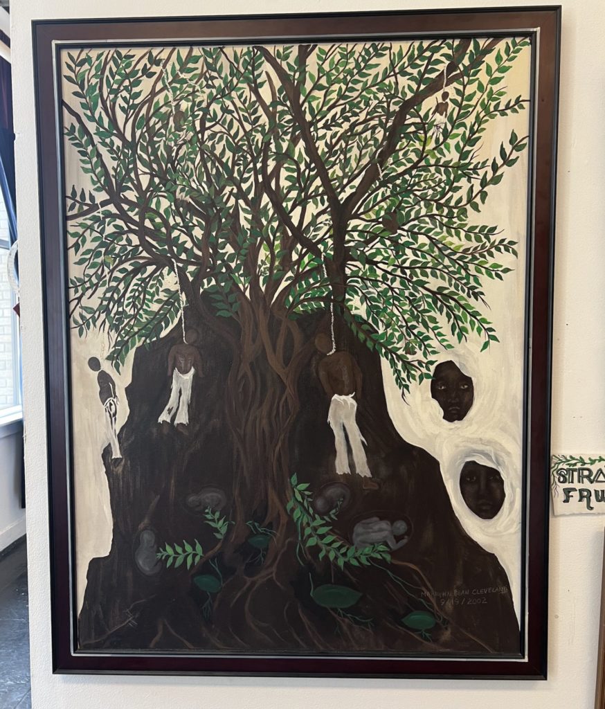 Painting by Marilyn Dean Cleveland. A large tree, the trunk is wider than the branches and takes up most of the frame- 2 black men are hanging from the tree in nooses.