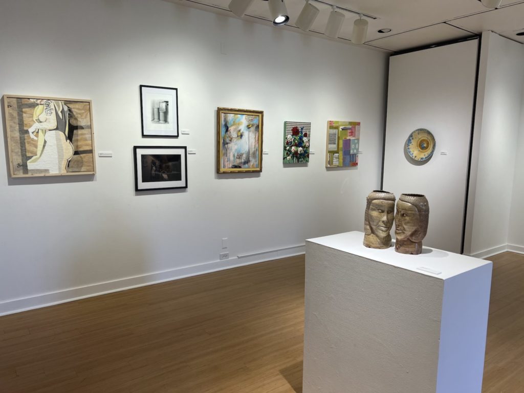 Art Around the Block gallery shot. On the far wall are six paintings. A duo of face sculptures sit on a large square white pedestal in the foreground.