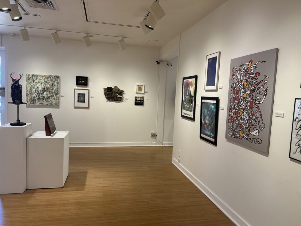 Gallery shot of Art Around the Block. Paintins are on the far back wall and right hand side; there are sculptures on the left center of the room on different heights of white square pedastals