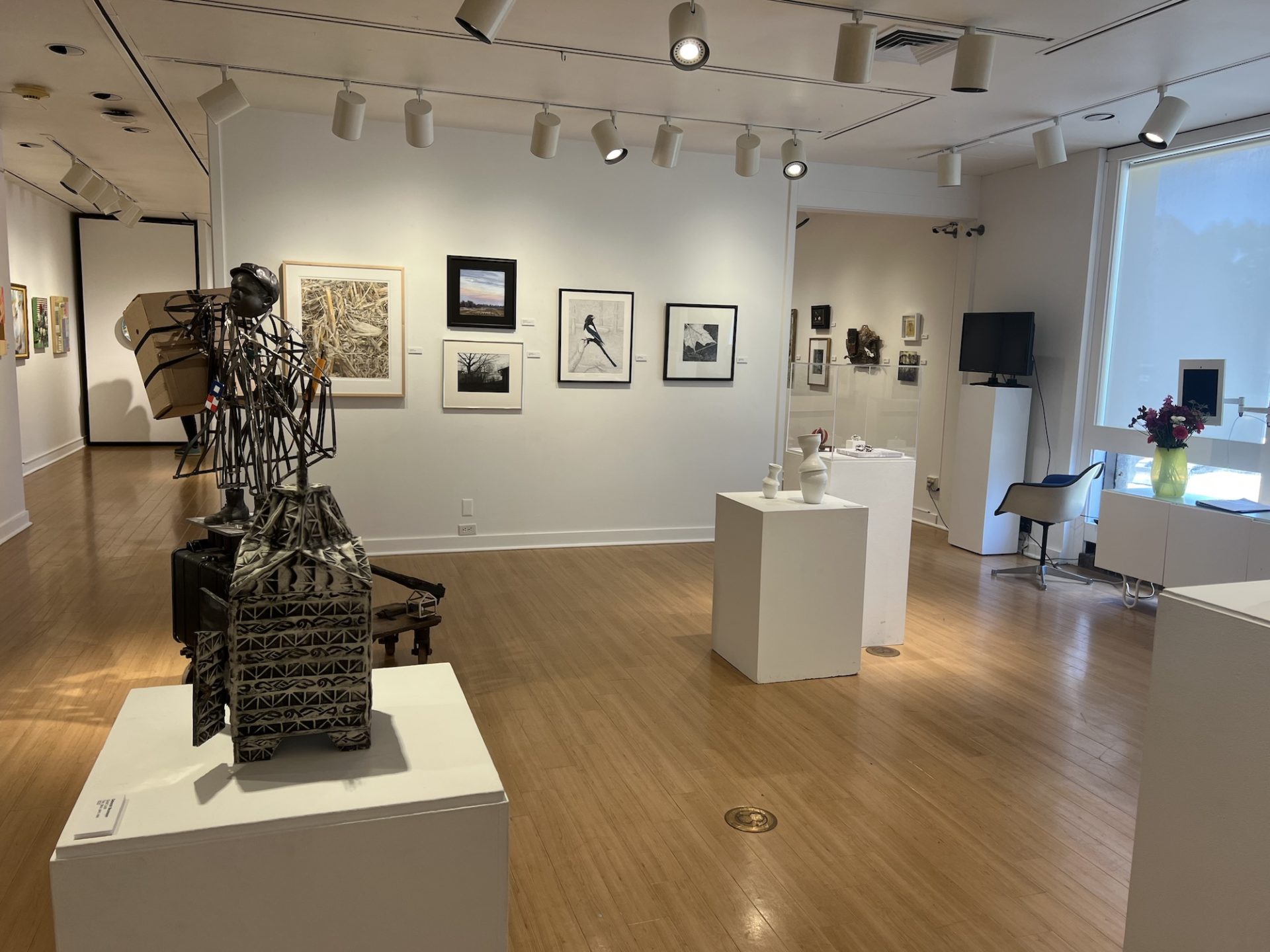 An overview shot of the art around the block gallery show. In the foreground are various sculptures on white square pedestals. On the far wall are small pieces of artwork. There is an open doorway on the right leading to the second half of the gallery.