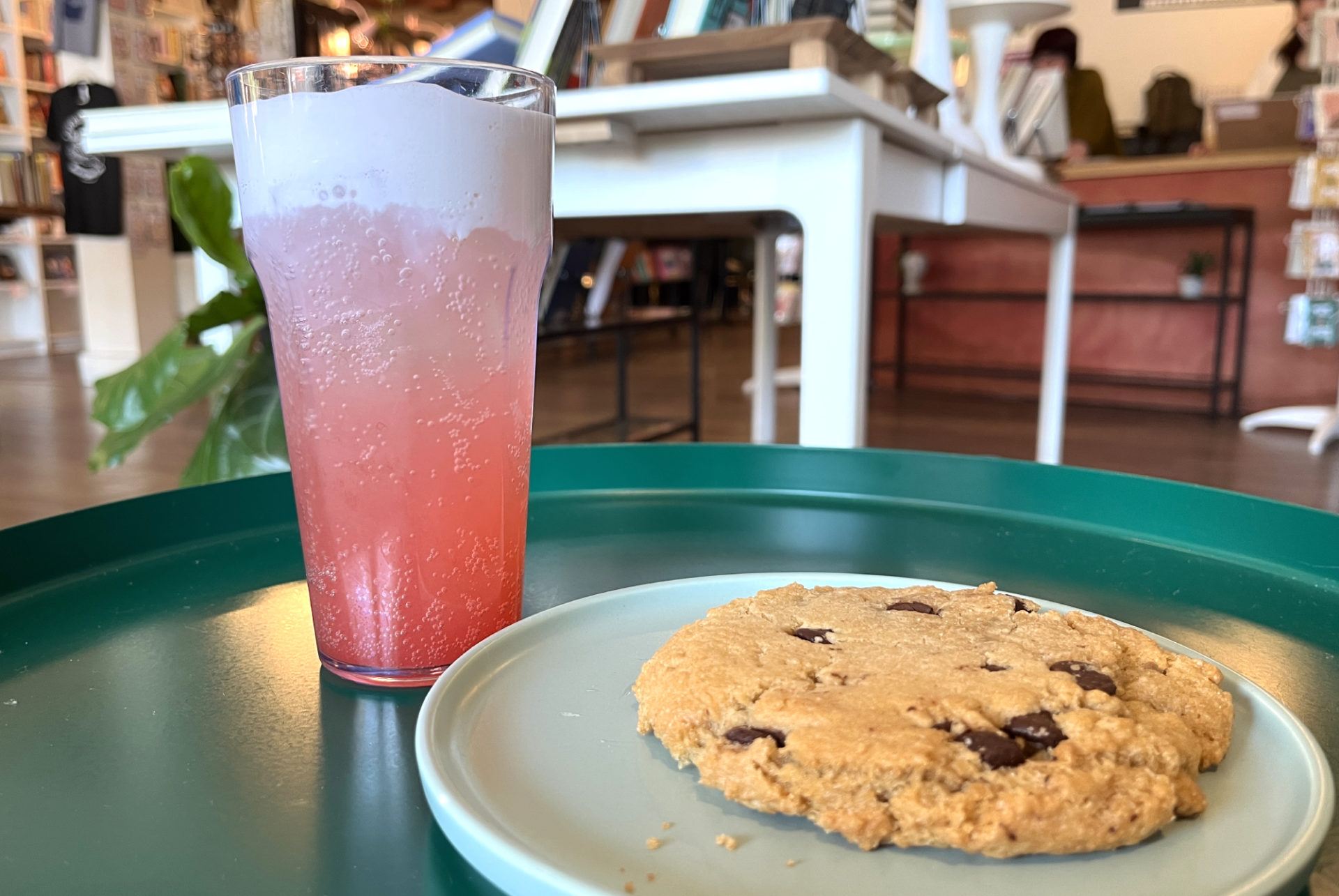 A tall glass of a pink beverage topped with cream and a light green plate with a chocolate chip cookie are both on a teal green side table at The Literary. Bookshelves are visible in the background.