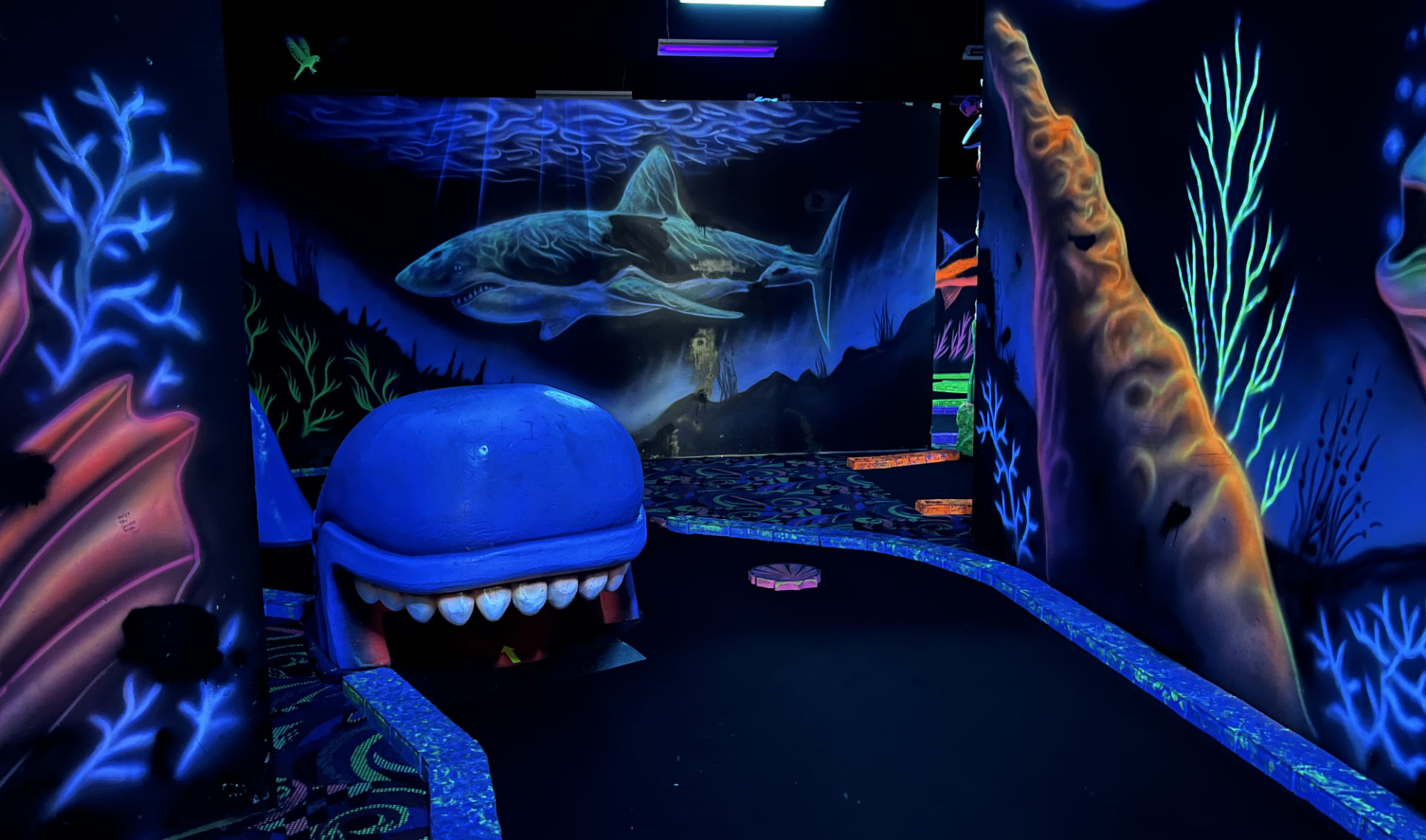 View of first hole of black light mini golf at Arrowhead Lanes. A black light-lit space shows under-the-sea murals on the walls featuring sea plants and sharks and fish. The mini golf hole features a sperm whale through which you are intended to hit the gold ball.