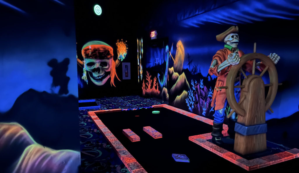 Black light mini golf hole at Arrowhead Lanes. The theme of the hole is a skeleton pirate ship, and there is a large sculpture of a skeleton at the wheel of a ship in the course. On the walls are paintings of shipwrecks and a large pirate skull.