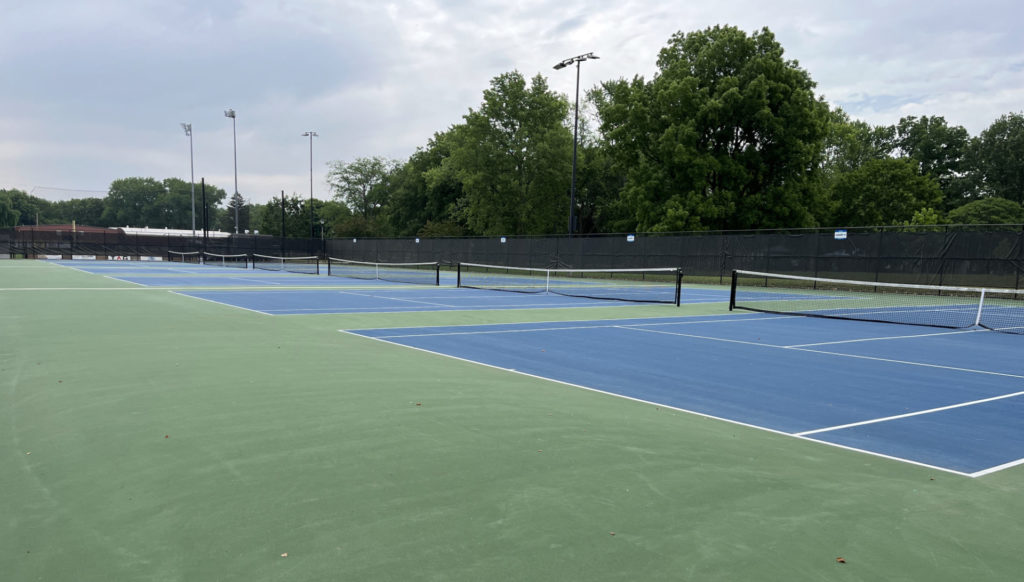 Six tennis courts, fenced in, at Spalding Park. The courts are blue with white lines, and the surround areas are green.