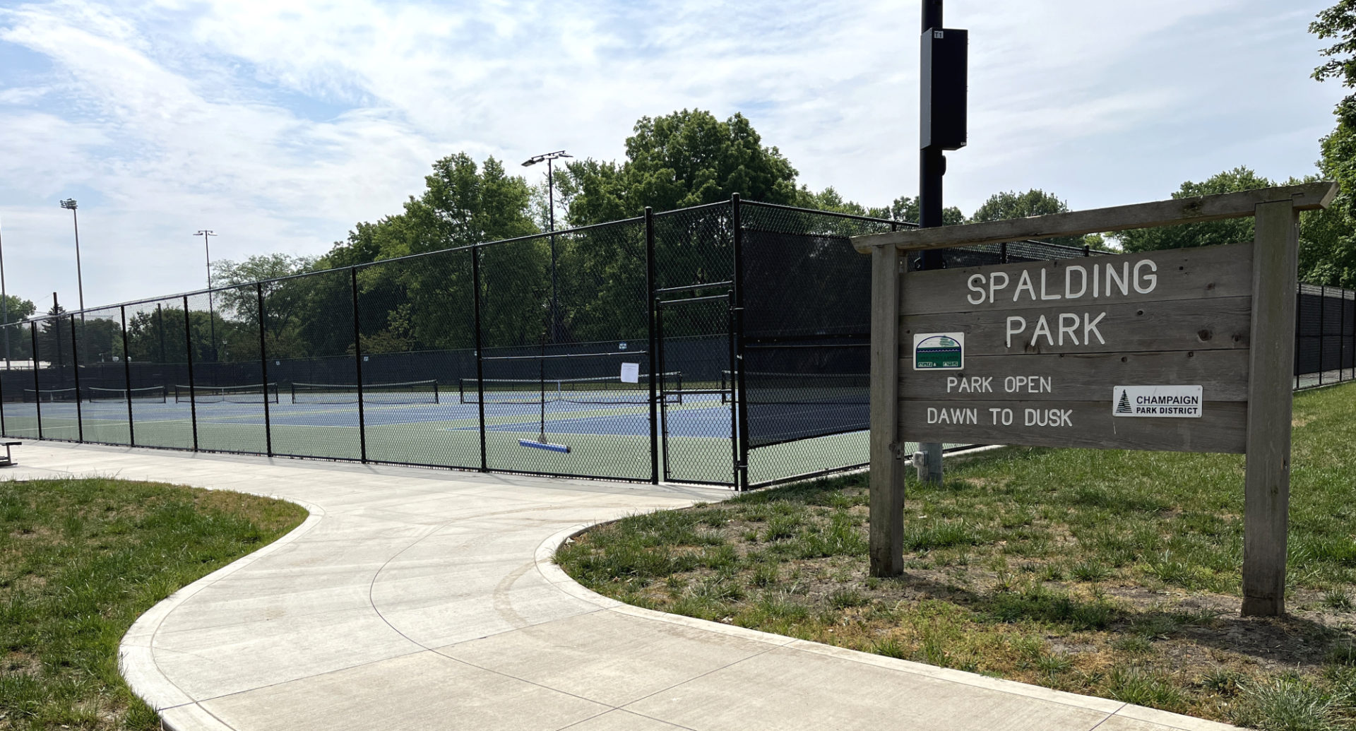 Large wood sign that reads Spalding Park is located near a sidewalk and tennis courts. The courts are to the left of the image.