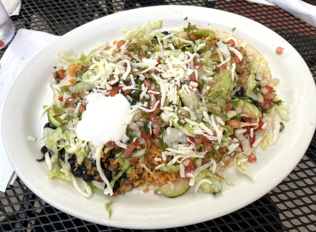 A white plate on an outdoor, black metal table. A huarache is on the plate and is covered in black beans, rice, lettuce, veggies, sour cream, and guacamole.
