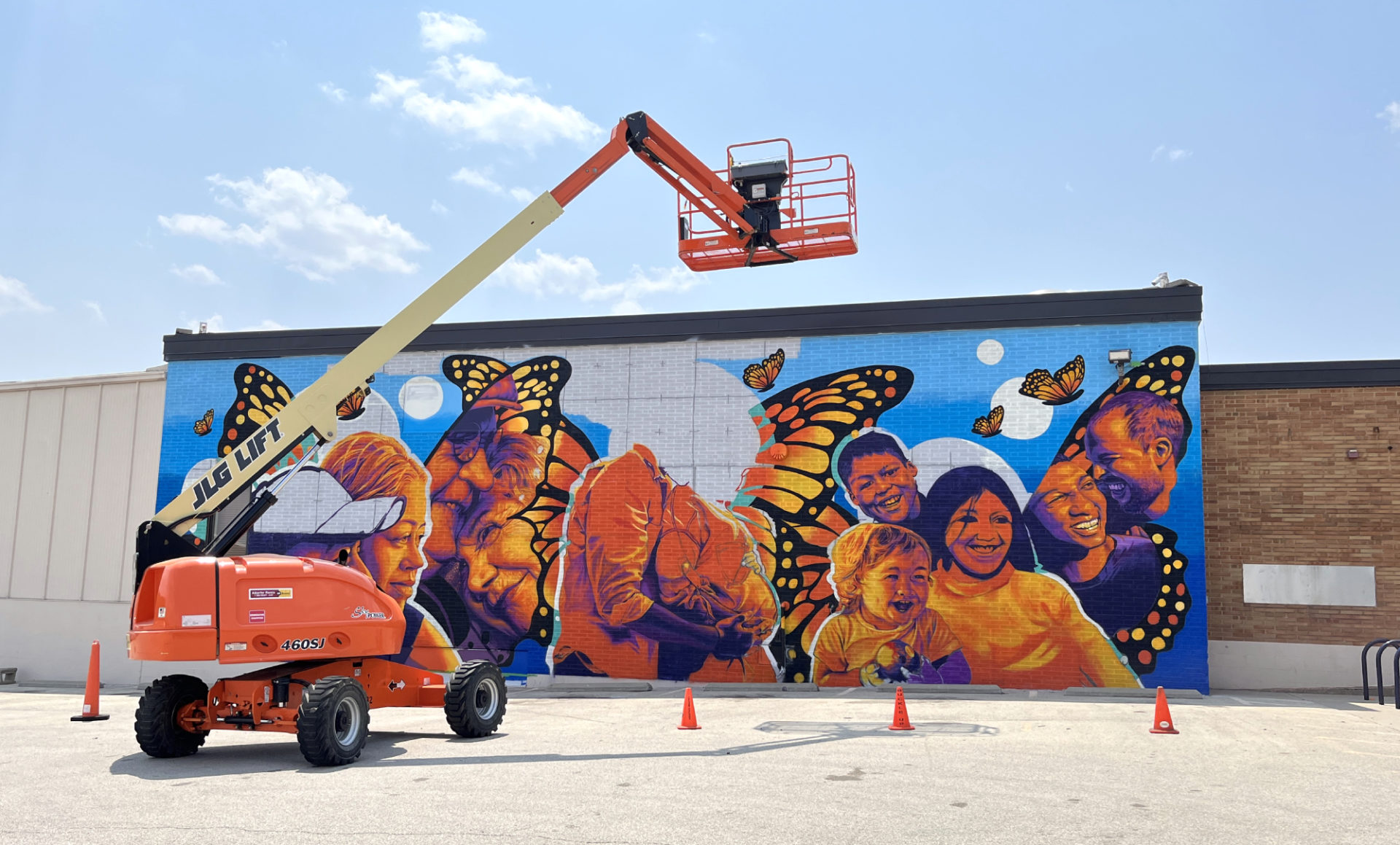 An in-progress mural at Champaign Urbana Public Health District by Jose Vazquez. It features 11 different people, all painted among and the colors of monarch butterflies. There are orange cones and a large cherry picker parked in front.