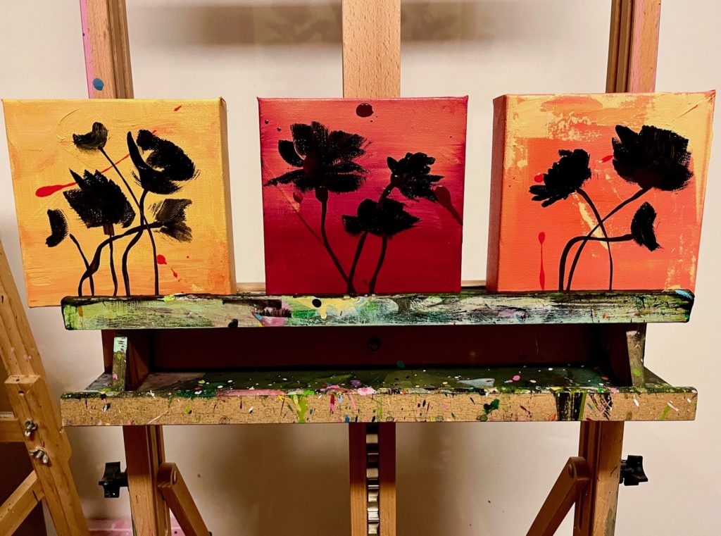 Three square canvases on an easel. The left canvas is primarily yellow, the middle is primarily red, the right primarily orange. All have black silhouettes of florals. 