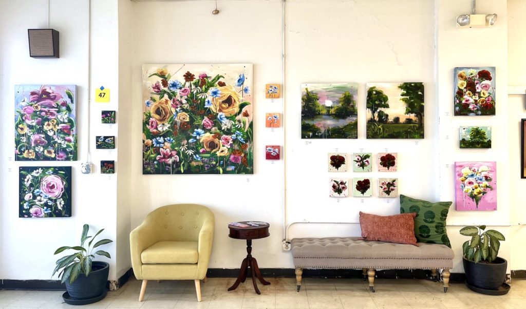Faith Gabel's display at Boneyard arts festival. Her floral paintings, mostly square canvases, are hanging on the wall. In front is a yellow velvet arm chair and tufted velvet bench with pillows. 