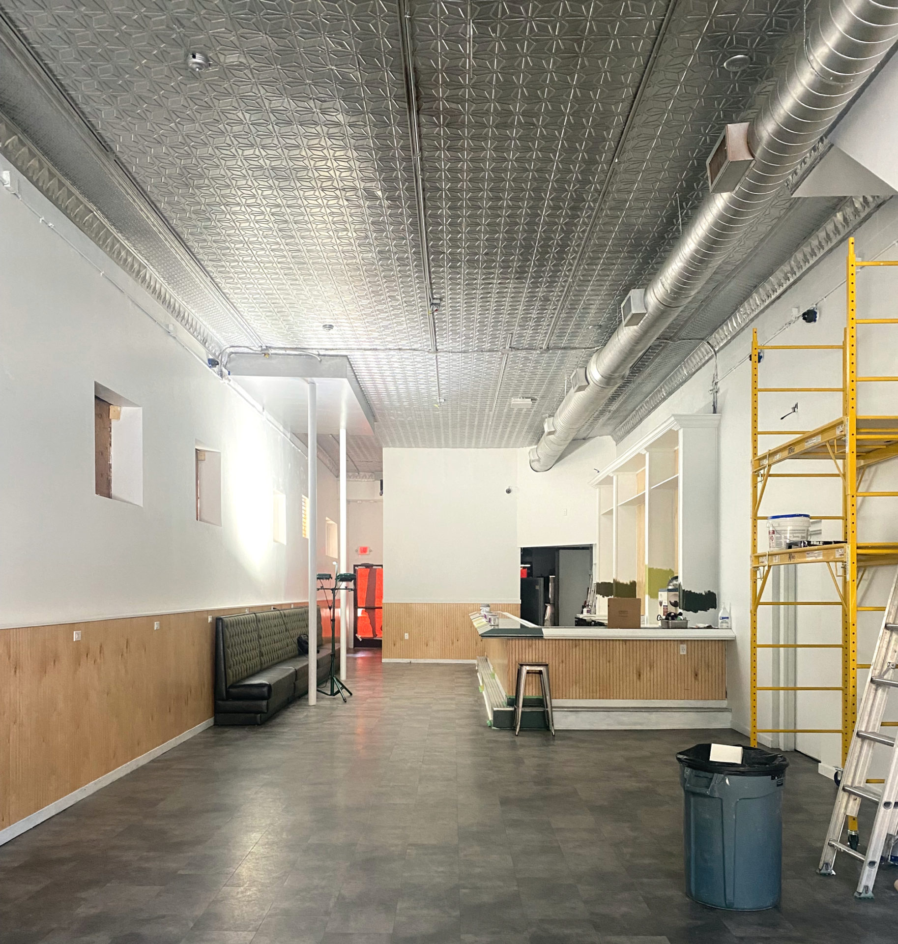 Interior of Gallery, an art bar in Urbana, under construction. The walls are white with light wood beadboard around the walls and bar. The floor is gray. There is a tin ceiling. Some ladders are stacked on the right side of the image, next to yellow scaffolding.