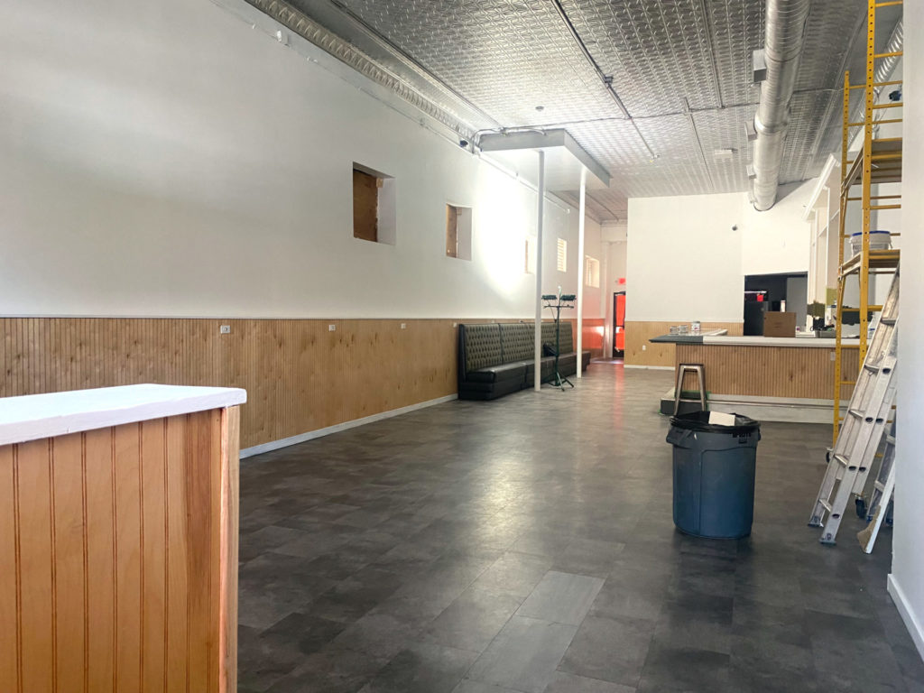 Interior of Gallery, an art bar in Urbana, under construction. The walls are white with light wood beadboard around the walls and bar. The floor is gray. There is a tin ceiling. Some ladders are stacked on the right side of the image.