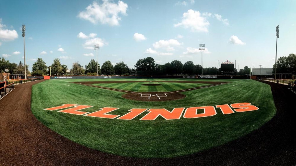 a wide angle view of the Illinois baseball field. There are freshly mowed grass lines the brown baseball diamond and the worlds "Illinois" in big orange block letters. Above there is a blue sky with white clouds. 