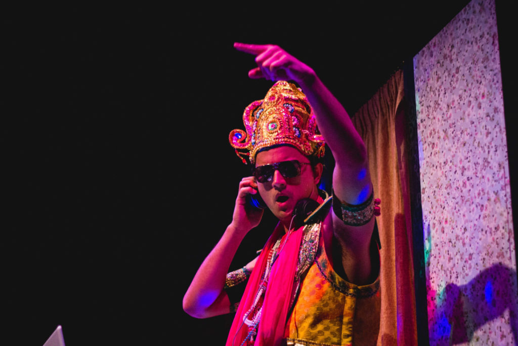 Indian Ink Theatre. An Indian man wearing a yellow elaborate traditional Indian outfit with a jeweled hat is Dj'ing. He has large headphones and one arm up in the air.