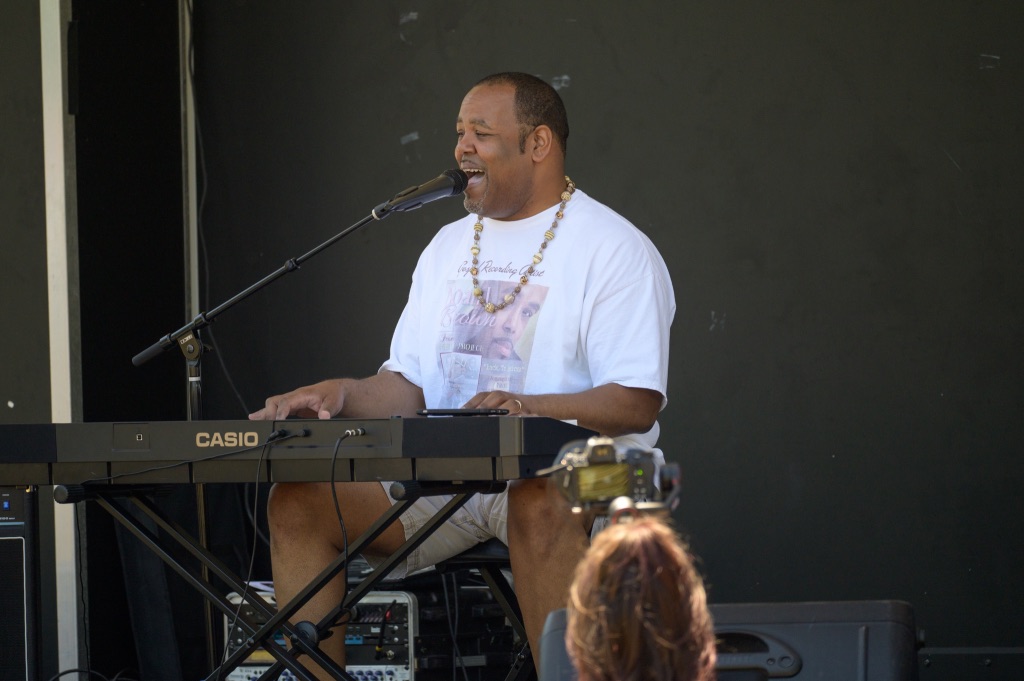 A Black man in a white t-shirt sits at a keyboard and sings into the attached microphone. There is a black wall behind him.