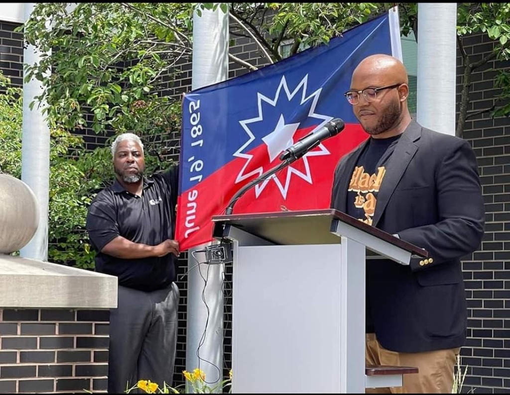 A Black man in a black shirt and jacket and tan pants stands at a podium. to the left of him another man in a black shirt and gray pants holds a red and blue flag with a white star and white outline on it. 