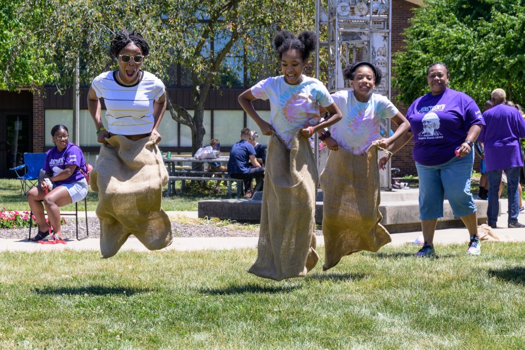 3 young Black girls are jumping off the ground in a sack race. They are laughing while spectators watch.