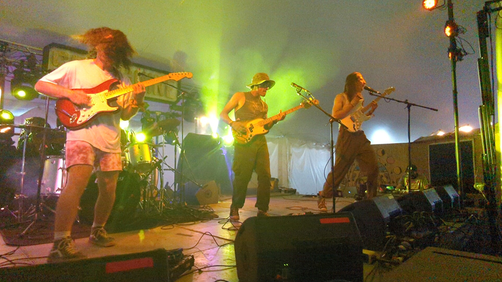 3 members of Kangaroo court on a stage lit with green light inside a tent. You can see 2 guitar players and a bass player.