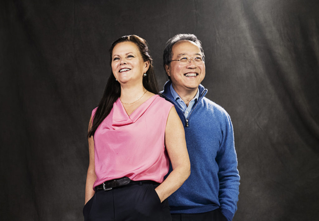 Kathryn Stott stands on the left; she has long dark hair and a pink sleeveless shirt, her hands are in the pockets of her black dress pants. Yo-Yo Ma stands slightly behind her, looking off camera to the right. They are both smiling. Ma wears a blue half-zip sweater. 