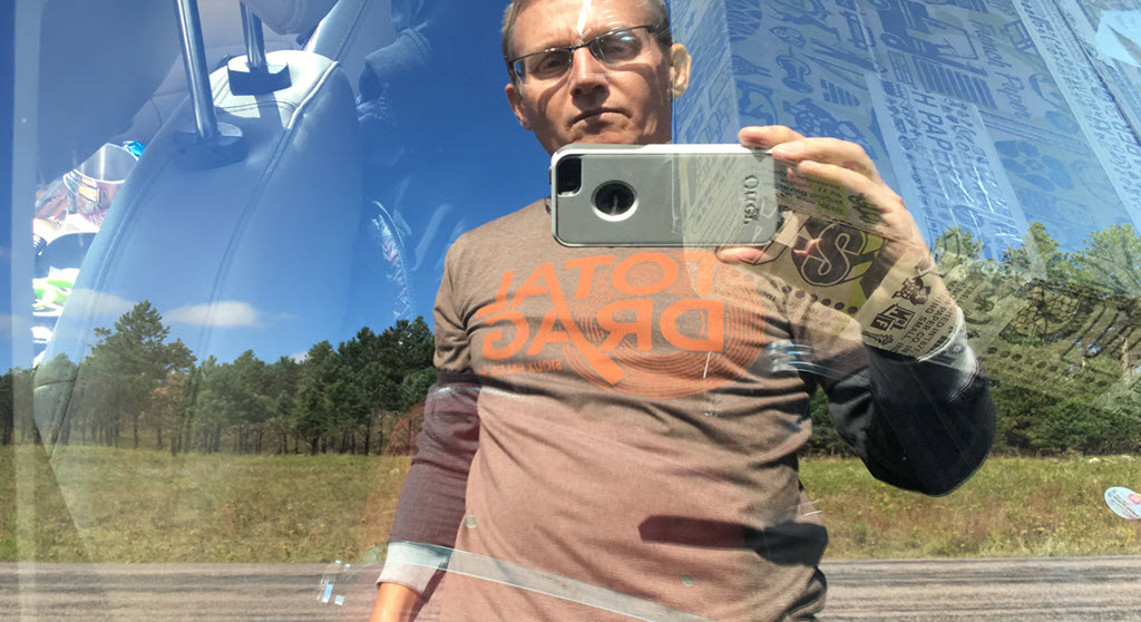 A man holds a cell phone camera up to the window of a car and takes his photo in the reflection on the window.
