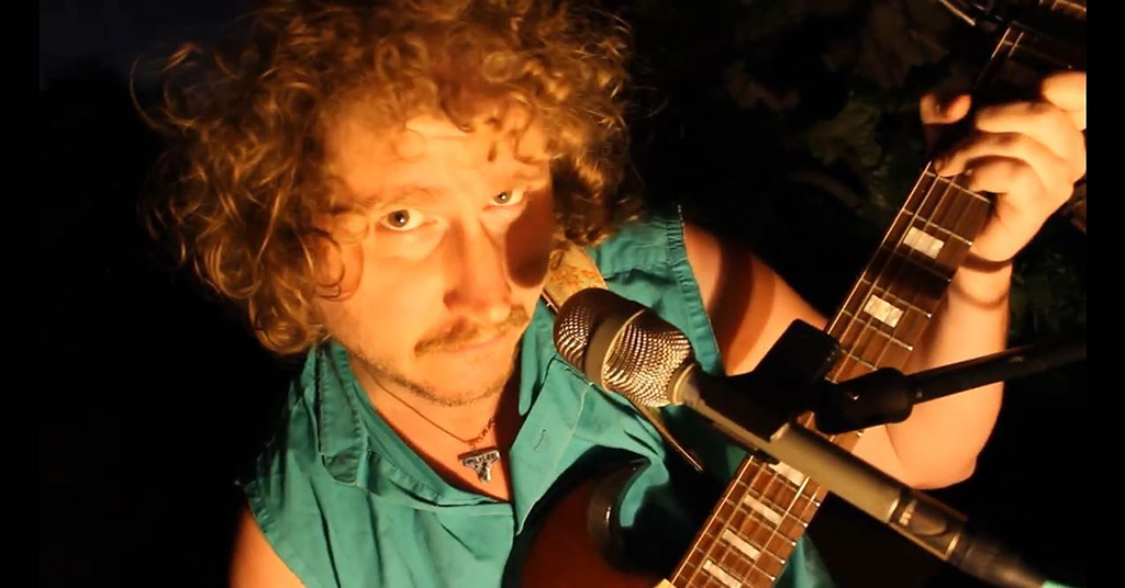 A man holding a guitar with a microphone to his face looking up at the camera. He has a turquoise sleeveless shirt, curly red hair and brown eyes.