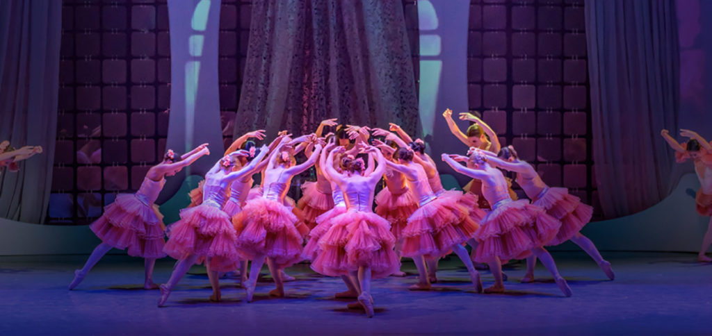 A large group of ballerinas wearing matching pink dresses with full multi-layered pink tutus are in a circle, arms extended above their heads, as they lean towards the center of the circle. A scene from the Nutracker ballet.