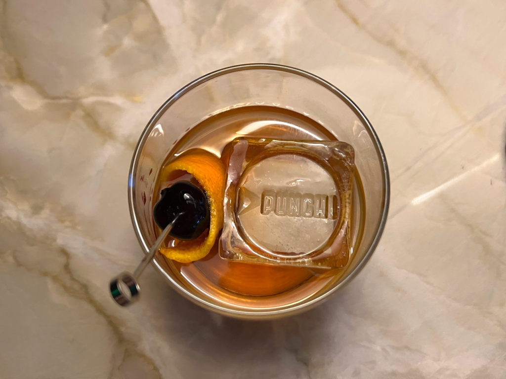Overhead shot of an old fashioned in a rocks glass with a cherry and orange peel garnish, and a cube of ice stamped with the word Punch.
