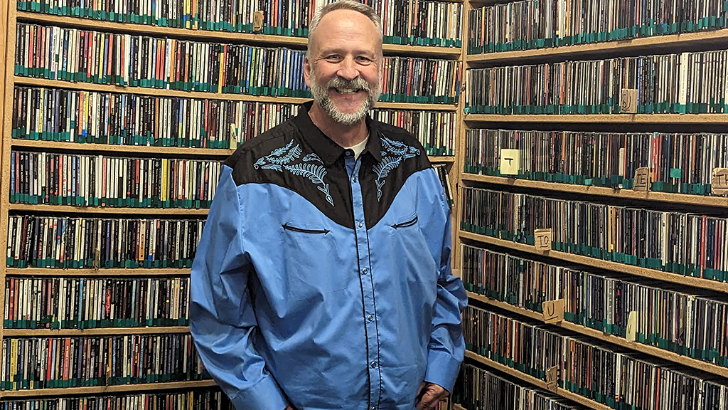 A man with a blue and black country western shirt in front of huge racks of CDs inside a radio station