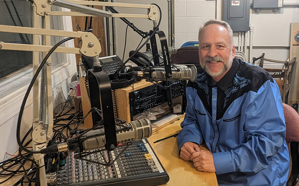 A man with a beard wearing a blue and black shirt in a radio studio in front of the broadcasting equipment