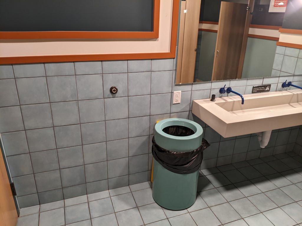 a bathroom sink surrounded by light blue tile and a orange and white painted mirror surround. There is also a teal garbage can. 