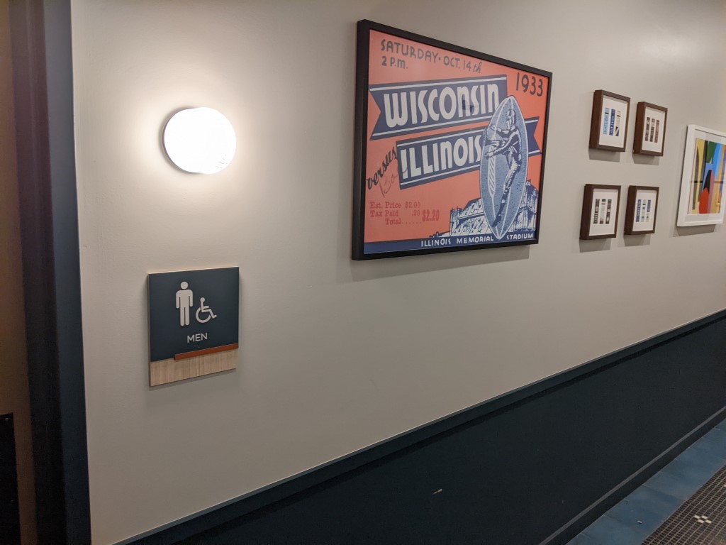A hallway with a wall painted white on top and blue on the bottom. with a bathroom plaque, a circular light, and an orange and blue framed print.