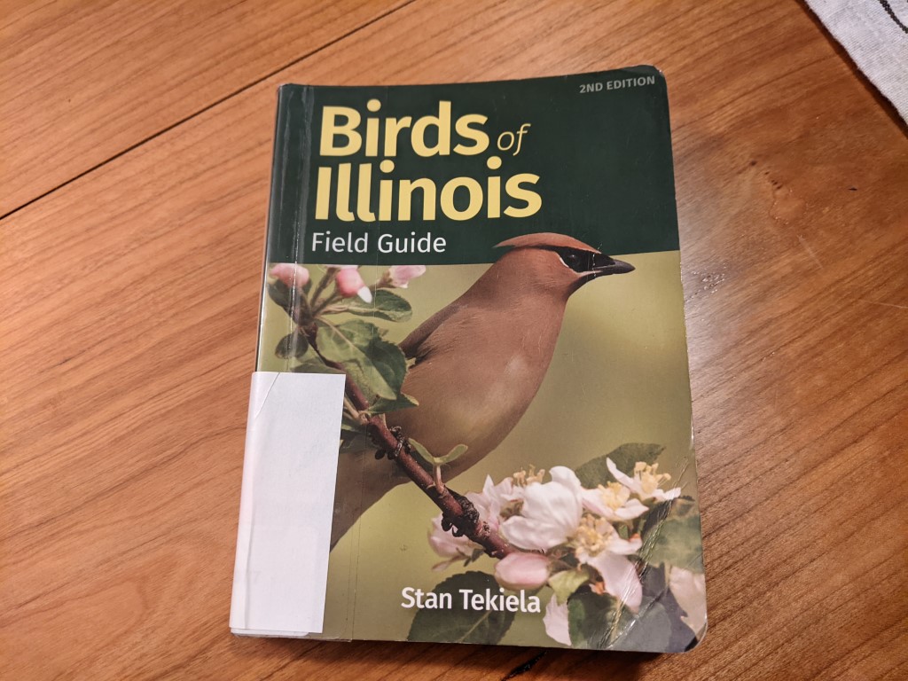 a picture of a book cover on a wooden table. The cover says "Birds of Illinois" with a brown bird sitting on a flowering branch. 