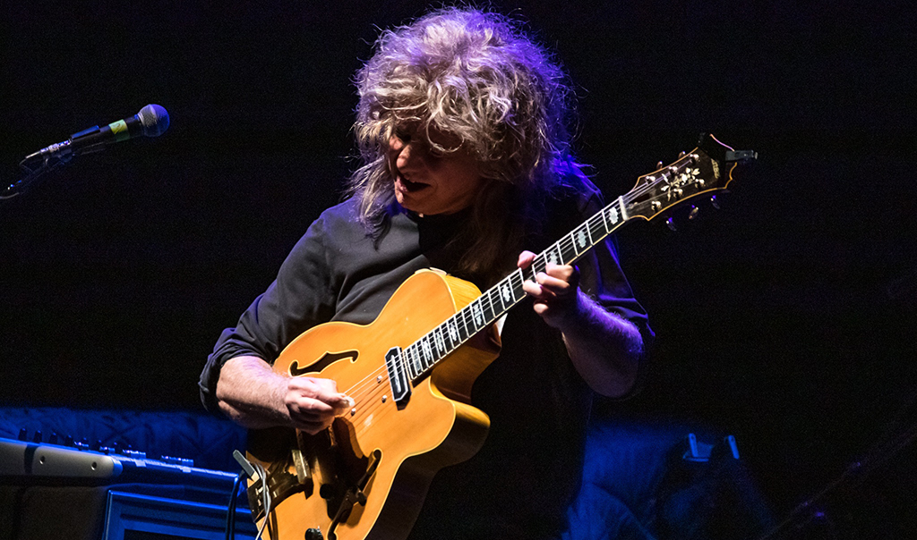 Closeup of musician Pat Metheny playing a guitar on stage. He's looking down at the stage while playing and the stage lighting is purple.