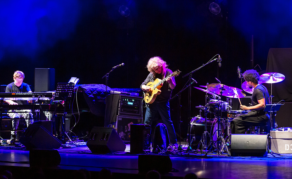 Musician Pat Metheny and two of his bandmates onstage peforming. The stage is bathed in purple light