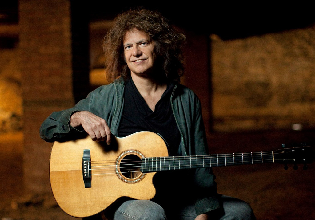 A man with very long and frizzy hair holding a guitar and sitting down in what looks like modern ruins