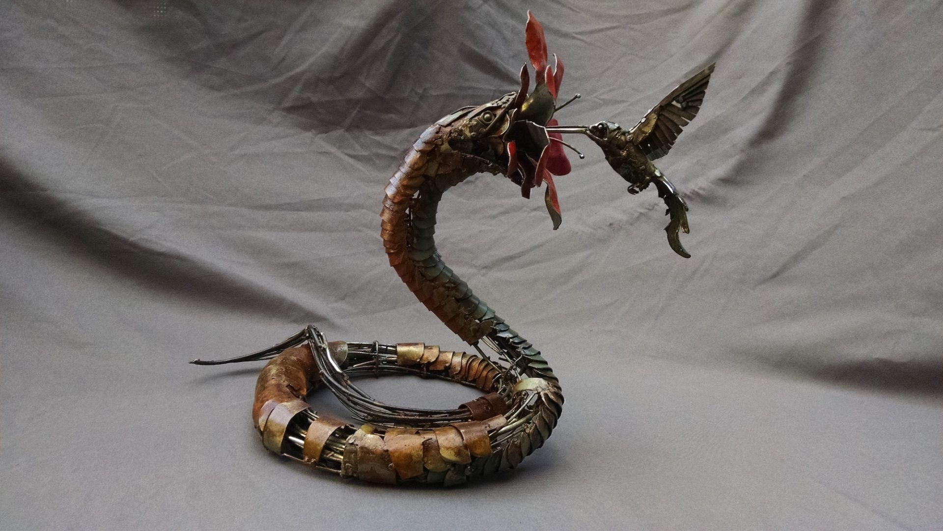 A metal sculpture by Andy White. A hummingbird attempts to pollinate a large flower which it cannot see is being held in the mouth of a large snake.
