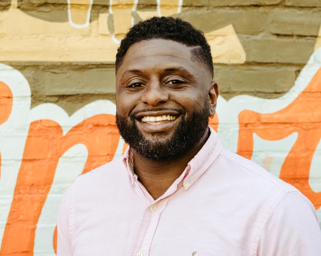 A black man with short dark hair and a dark beard is wearing a pink button up shirt and standing in front of a brick wall with a an orange and brown mural painted on it. 