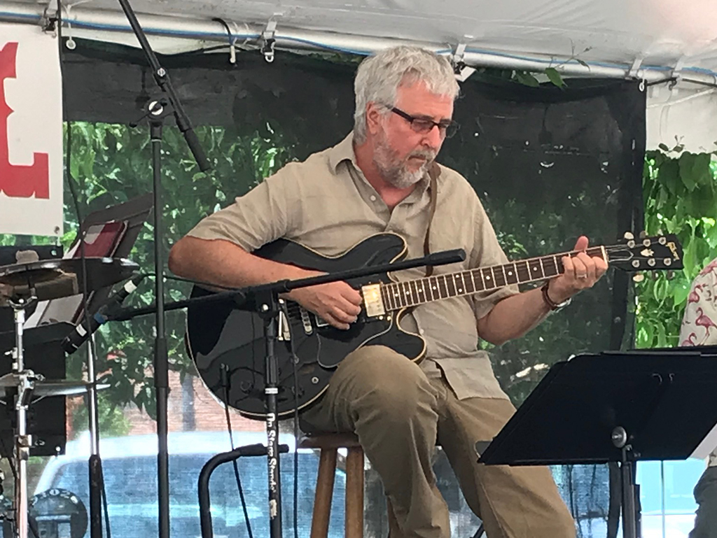A man in a tan shirt and pants sitting down on an outdoor stage playing a black guitar.