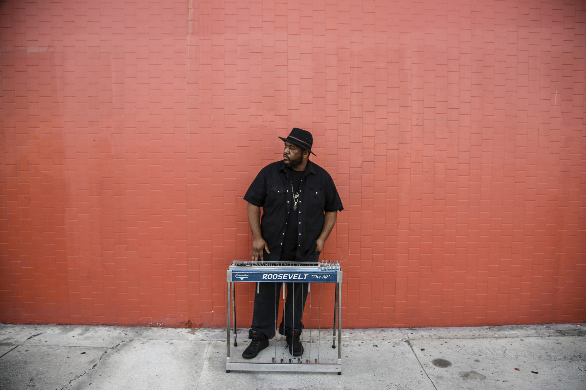 Roosevelt Collier, a black man, is standing against an orange-red brick wall outside. He is wearing all black and a black hat. He is looking off camera while a keyboard is in front of him.