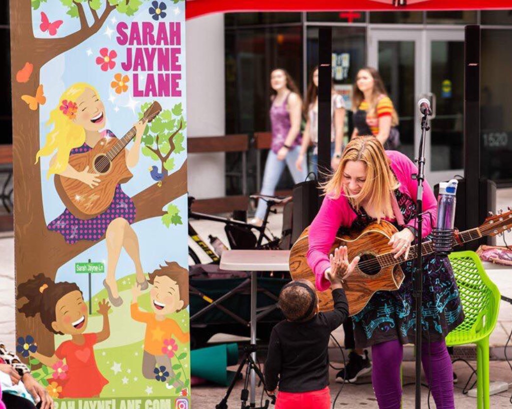 A white woman in a pink cardigan, a music note printed dress, and purple pants is leaning over her guitar to high five a small child with a black shirt and red pants. To the left of her is a large sign that says Sarah Jayne Lane